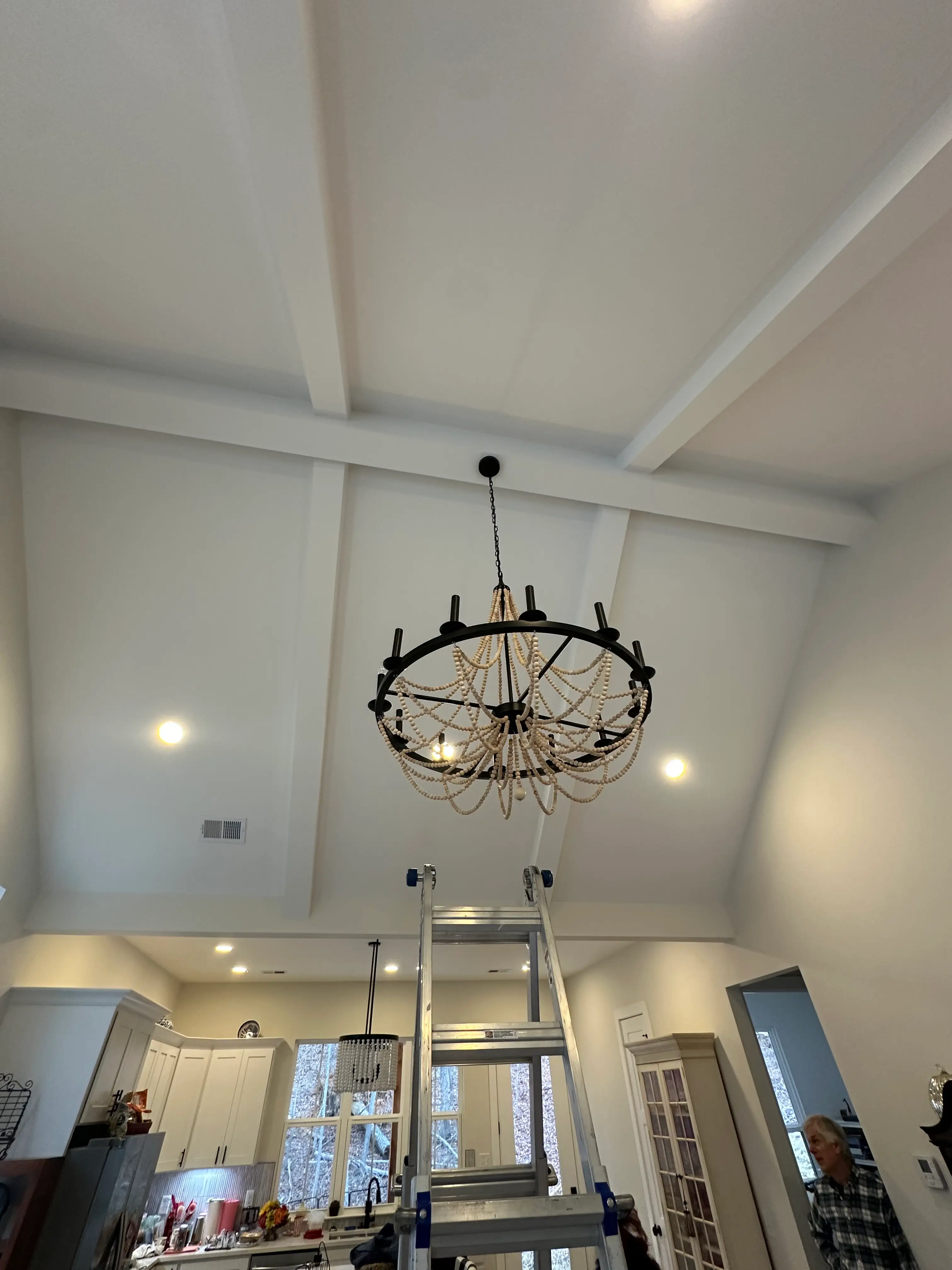 Black and wood-bead chandelier hanging from a tall white coffered ceiling above a ladder